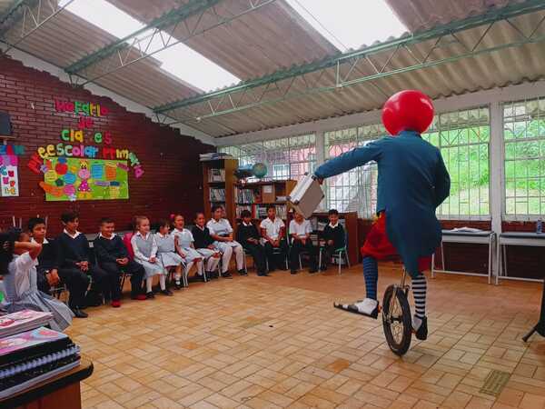 Payaso en monociclo en aula con niños en el fondo.